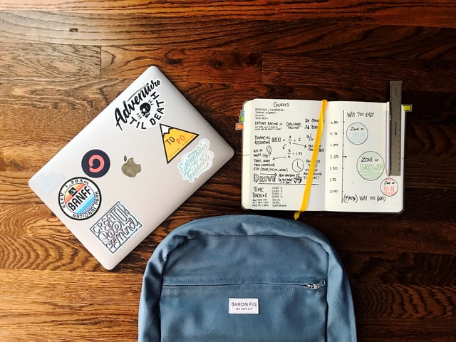 closed laptop, open notebook, and a blue backpack resting on a brown table